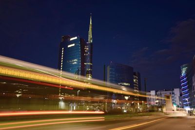 Light trails on road by buildings against sky at night