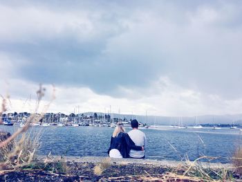 Rear view of young couple with arm around sitting by sea 