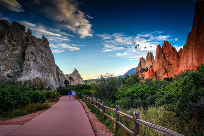 Rear view of people walking on footpath by rock formation and plants against sky at colorado springs