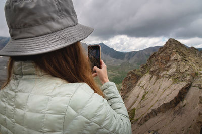 Woman in panama stands with her back to the camera and photographs the mountains on the phone, a