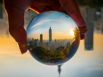 Cropped hand holding crystal ball with reflection of taipei 101 in city during sunset