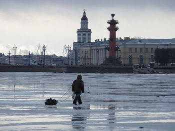 Full length of fisherman walking on snow against buildings in city