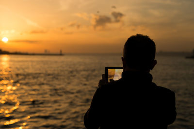 Rear view of silhouette boy photographing sea at sunset