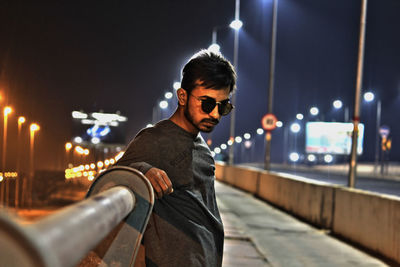 Portrait of young man wearing sunglasses while standing by railing at night