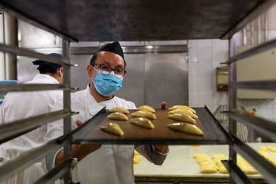 Male chef wearing uniform and medical mask preparing tasty pastry in kitchen of bakery during coronavirus epidemic