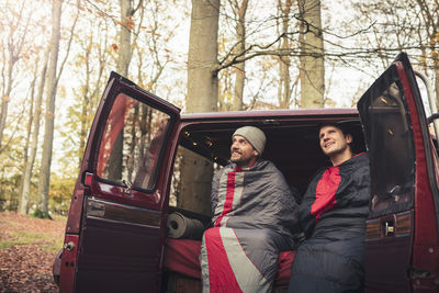 Smiling male friends under blankets sitting in camping van