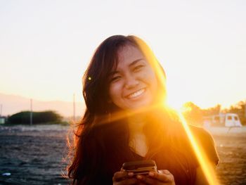 Portrait of happy young woman using smart phone against clear sky during sunset