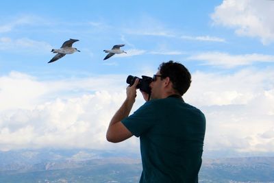 Rear view of young man photographing seagulls flying in sky