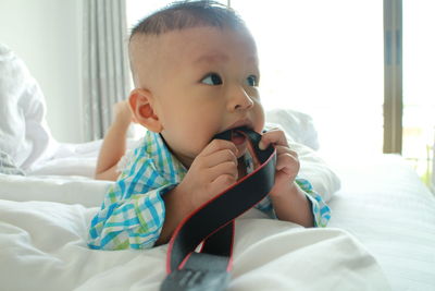 Close-up of thoughtful baby boy biting strap while lying on bed at home