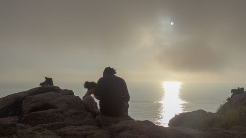 Rear view of man sitting on rock by sea against sky