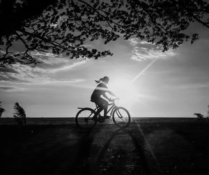 Side view of silhouette woman riding bicycle by sea against sky during sunny day