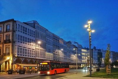 Illuminated street by buildings against sky at night