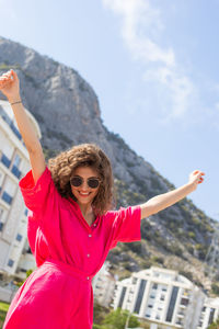 Portrait of young woman with arms raised standing against sky