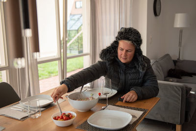 A woman sits at the dining table, dressed warmly, and eats