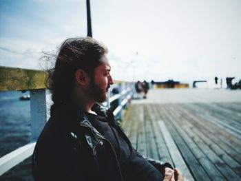 Young man looking away while sitting by railing against sky