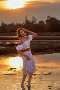 Full length of young woman standing at beach against sky during sunset