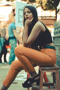 Model sitting on a wooden bank wearing black top and yellow pant and black shoes looking at camera