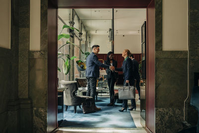 Full length of male and female professionals doing handshake while standing by colleague in hotel lounge