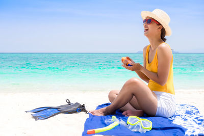 Happy young woman applying suntan lotion while sitting on beach against sky
