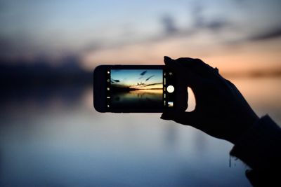 Cropped person photographing lake with mobile phone during sunset