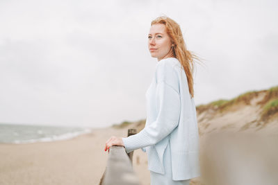 Portrait of young red haired woman in light blue sweater on sand beach by sea in storm