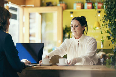 Barista serving coffee to woman using laptop in cafe