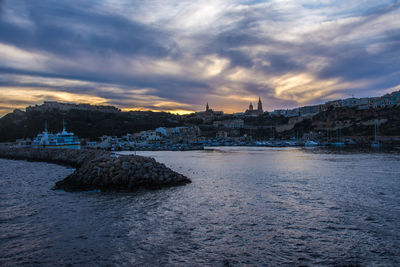 Buildings by sea against cloudy sky at sunset