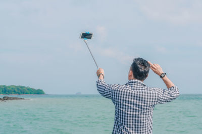 Rear view of man taking selfie from mobile phone by sea