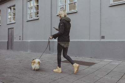 Full length of blond woman walking with pomeranian on footpath by building in city