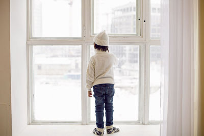 Boy in a white sweater and hat stands rear at the window in winter in large apartment on christmas