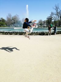 Woman jumping over walkway against fountain during sunny day