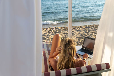 Blonde woman using and typing on laptop computer screen while laying down on daybed on the beach