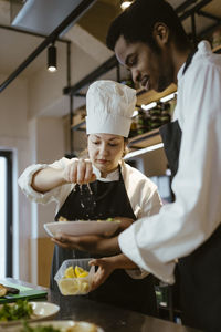 Female chef squeezing lemon on salad bowl held by male colleague in commercial kitchen