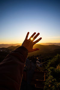 Cropped hand of person against mountains and sky during sunset