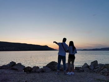 Rear view of man and girl pointing while standing at beach against sky during sunset