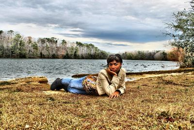 Portrait of woman lying on lakeshore against cloudy sky