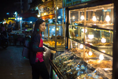 Side view of woman standing by illuminated store at night