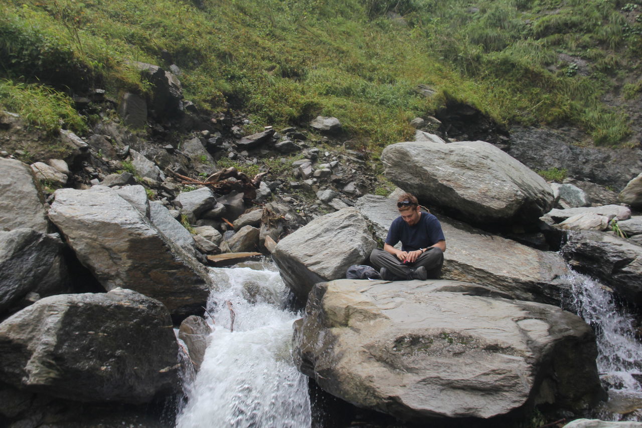 REAR VIEW OF MAN SITTING ON ROCKS BY WATERFALL