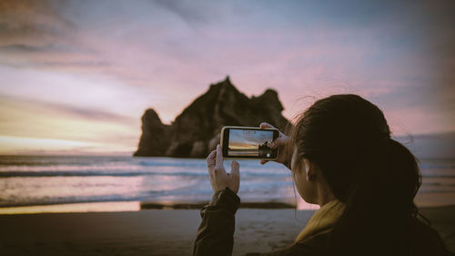 Side view of woman photographing against sky during sunset in wharariki beach