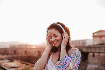 Smiling woman listening to music while standing against sky