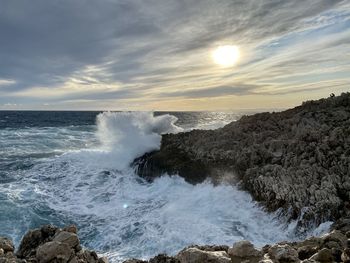 Waves splashing on rocks at shore against sky