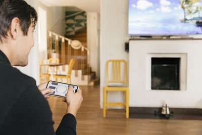 Man using smart home application on mobile phone while looking at television in living room at home