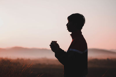 Side view of silhouette man having drink while standing against clear sky during sunset