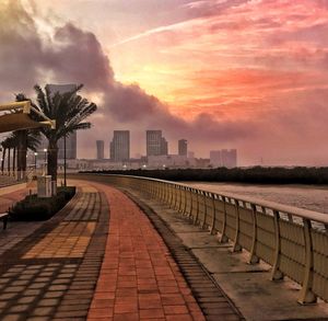 Footpath by palm trees and buildings against sky during sunset