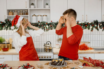 Playful positive 
husband and wife having fun in the kitchen covering eyes with gingerbread cookies
