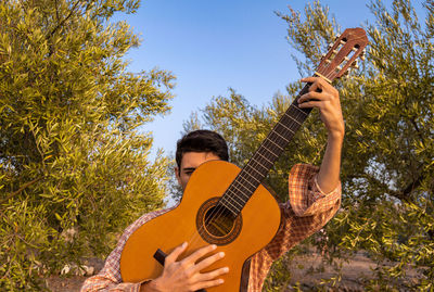 Young man holding guitar against trees