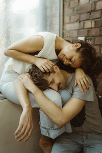 Smiling man with eyes closed resting on girlfriend's lap while sitting at home