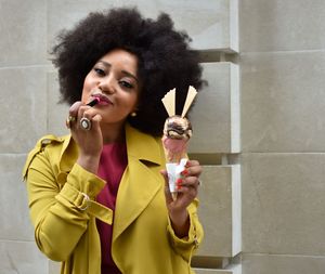 Portrait of woman applying lipstick while holding ice cream against wall