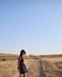 Woman standing on dirt road amidst grassy landscape against clear sky