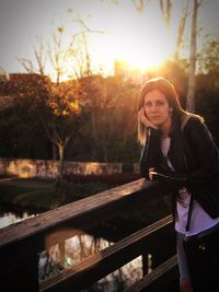 Portrait of young woman standing on railing against river at sunset